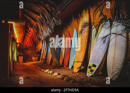 Eine Sammlung verschiedener farbenfroher Surfbretter, die als Dekoration neben einem Gehweg verwendet werden und auf einer Wand ruhen. Das Foto wurde in der Nacht mit einem Haufen von aufgenommen Stockfoto