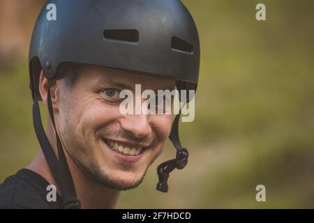 Das junge kaukasische männliche Model trägt einen schwarzen Fahrrad- oder Skate-Helm und lächelt. Cooler Typ, der draußen einen Helm trägt Stockfoto