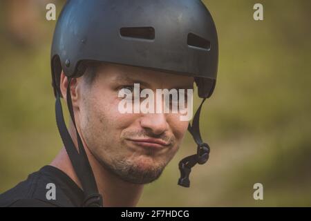 Das junge kaukasische männliche Model trägt einen schwarzen Fahrrad- oder Skate-Helm und macht ein Entengesicht. Cooler Typ, der draußen einen Helm trägt Stockfoto