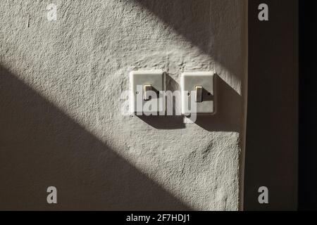 Ein paar alte europäische elektrische Schalter, die an einer Wand unter einem starken Seitenlicht montiert sind. Sichtbare alte Knete oder Gips an der Wand. Stockfoto