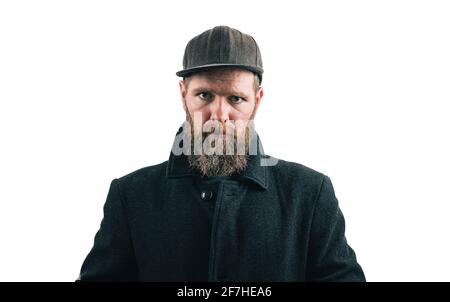 Wütend aussehender Hipster-Mann mit Bart und Baseballmütze, der einen schwarzen Wintermantel auf weißem Studiohintergrund trägt. Wütender bärtiger Mann. Stockfoto