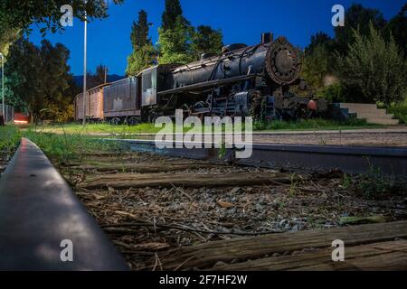 Alte Dampfeisenbahn in der Nacht geparkt als Außenausstellung von Eisenbahnen in einem Park in der griechischen Stadt Kalamata. Dampflokomotive mit Wagen in Par Stockfoto