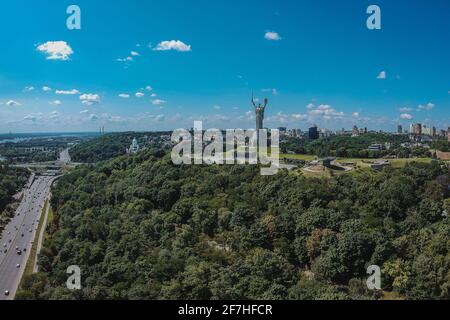 Panorama des Mutterland-Denkmals in Kiew, das an einem sonnigen Tag mit blauem Himmel aus dem Wald ragt. Majestätisches Foto einer großen Statue auf dem Hügel in Kvyv, U Stockfoto