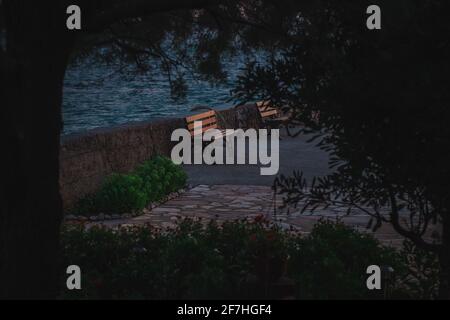 Einsame Bank am Rande eines Strandes am Abend. Holzstrand an einem Strand, der von Büschen und Bäumen umgeben ist. Stockfoto