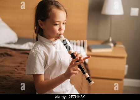 Seitenansicht des talentierten entzückenden kleinen Mädchens, das Flöte spielt und im Schlafzimmer auf dem Bett sitzt und wegschaut. Stockfoto