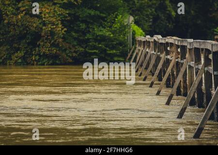 Überflutete Holzbrücke mit Straße, aufgrund von starkem Regen und schlechtem Wetter. Schmutziges Flusswasser fließt direkt über die Holzbrücke. Gefährlich Stockfoto