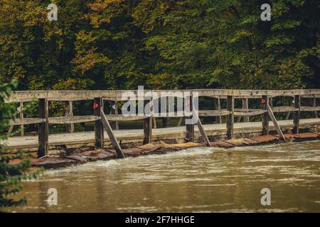 Überflutete Holzbrücke mit Straße, aufgrund von starkem Regen und schlechtem Wetter. Schmutziges Flusswasser fließt direkt über die Holzbrücke. Gefährlich Stockfoto