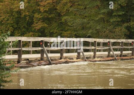 Überflutete Holzbrücke mit Straße, aufgrund von starkem Regen und schlechtem Wetter. Schmutziges Flusswasser fließt direkt über die Holzbrücke. Gefährlich Stockfoto