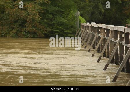 Überflutete Holzbrücke mit Straße, aufgrund von starkem Regen und schlechtem Wetter. Schmutziges Flusswasser fließt direkt über die Holzbrücke. Gefährlich Stockfoto