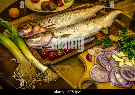 Zwei geschälte pollock-Fische, gefüllt mit Kräutern, Zitronenkeilen mit Gewürzen auf einem Tablett neben Lauch, Tomaten und Scheiben Zwiebel und grünem Basilikum Stockfoto