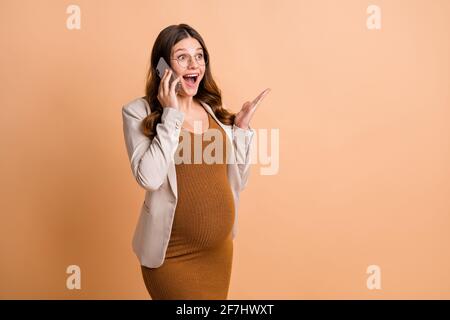 Foto von süßen hübschen Frau erwartet Kind tragen Jacke Arm Bauch reden moderne Gerät leeren Raum isoliert beige Farbe Hintergrund Stockfoto