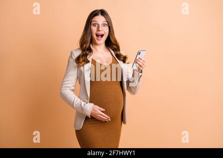 Foto von glänzend aufgeregt Dame wartet Baby gekleidet braunes Kleid Arm Bauch moderne Gadget isoliert beige Farbe Hintergrund Stockfoto