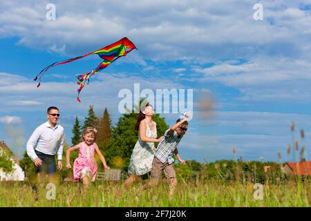 Glückliche Familie - Mutter, Vater, Kinder - die im Sommer über eine grüne Wiese laufen; sie fliegen einen Drachen Stockfoto