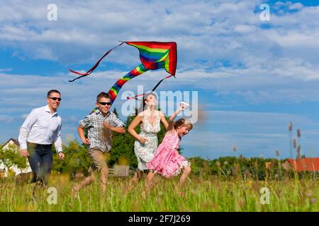 Glückliche Familie - Mutter, Vater, Kinder - die im Sommer über eine grüne Wiese laufen; sie fliegen einen Drachen Stockfoto