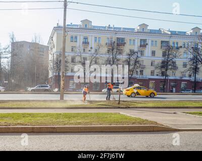Moskau. Russland. 06. April 2021. Orangefarbene uniformierte Hausmeister, die auf einer Straße in der Stadt den Rasen fegen. Sonniger Frühlingstag. Die Arbeit der Gemeinde Stockfoto
