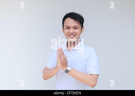 Junger asiatischer Mann in weißem Hemd geben Namaste Grußhände. Indonesischer Mann auf grauem Hintergrund. Stockfoto