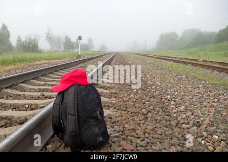 Schwarzer geschlossener Rucksack mit roter Baseballmütze auf der Oberseite Es steht auf dem braunen Granitschindel neben dem Schienen und Schwellen der Bahn gegen eine Stockfoto