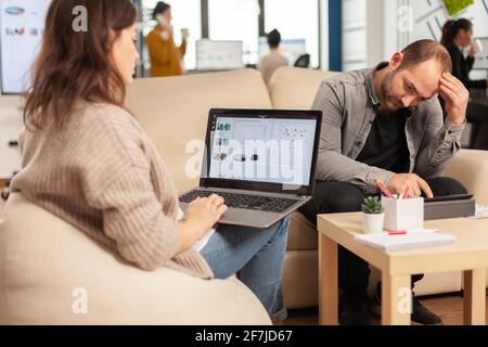 Rückansicht Video von jungen Geschäftsfrau sitzen auf Couch im Büro mit Laptop-Computer mit Grafiken, während Kollegen arbeiten im Hintergrund. Multiethnische Mitarbeiter planen ein neues Finanzprojekt Stockfoto