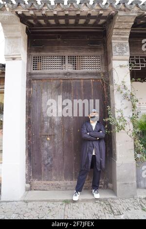 Ein asiatischer junger Mann, der sich in Schwarz mit schwarzem Hut und Gesichtsmaske kleiden und vor einem alten Haus mit Holztür steht. Stockfoto