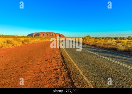 Straße, die zum Uluru Ayers Rock bei lebendiger Farbe des Sonnenuntergangs führt. Die riesige Monolith-Ikone des australischen Outbaks im Uluru-Kata Tjuta National Park Stockfoto