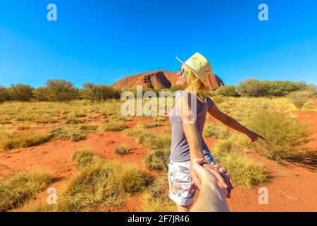 Folgen Sie mir, eine fröhliche Lifestyle-Frau, die am Ayers Rock im Uluru-Kata Tjuta NP die Hände hält. Konzept der Reise, bei der der Mann in der Hand im australischen Outback gehalten wird Stockfoto