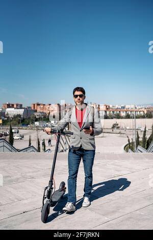 Stock Foto von einem Geschäftsmann in Jacke und Sonnenbrille Mit Rucksack auf Elektroroller und mit Smartphone stehen Stockfoto