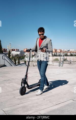 Stock Foto von einem Geschäftsmann in Jacke und Sonnenbrille Mit Rucksack auf Elektroroller und mit Smartphone stehen Stockfoto