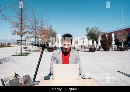 Junger Unternehmer, der draußen am Computer arbeitet und einen Kaffee nimmt Mit einem Elektroroller, der neben ihm steht Stockfoto