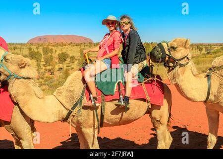 Paar Kamelreiten in der australischen Wüste des Northern Territory mit Uluru in der Ferne. Beliebte Aktivität, um den Monolith vom Kamel zu bewundern Stockfoto