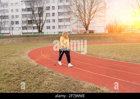 Eine ältere Frau in einer gelben Sportjacke praktiziert Nordic Walking im Freien auf dem Gummi-Laufband des Stadions. Ein sonniger Sonnenuntergang. Ältere Frauen gehen mit Stockfoto