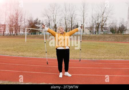 Eine ältere Frau in einer gelben Sportjacke praktiziert Nordic Walking im Freien auf dem Gummi-Laufband des Stadions. Ein sonniger Sonnenuntergang. Ältere Frauen gehen mit Stockfoto