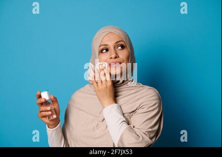 Schöne muslimische Frau mit bedecktem Kopf in Hijab, die mizellare Lotion aufwendet und Make-up von ihren Lippen entfernt. Make-up-Entfernungskonzepte auf blauem Backgr Stockfoto