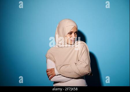 Junge Frau mit orientalischem Aussehen in beigefarbenem Hijab, die auf blauem Hintergrund posiert. Speicherplatz kopieren. Arabischer Mode-Style Stockfoto