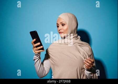 Junge Frau mit orientalischem Aussehen in beigefarbenem Hijab, die ein Smartphone in der Hand hält, isoliert auf blauem Hintergrund mit Kopierraum Stockfoto