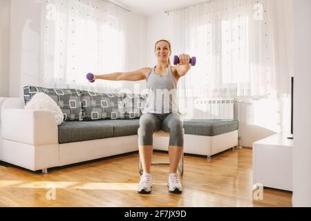 Lächelnd muskulöse Sportlerin sitzt auf dem Stuhl in ihrer Wohnung und macht Fitness-Übungen für Bizeps mit Hanteln. Stockfoto