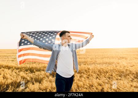 Junger Mann mit amerikanischer Flagge, während er auf dem Weizenfeld steht Vor blauem Himmel Hintergrund Stockfoto