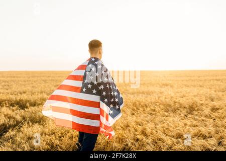 Junger Mann mit amerikanischer Flagge, während er auf dem Weizenfeld steht Vor blauem Himmel Hintergrund Stockfoto