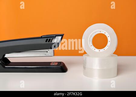 Hefter und Klebeband auf weißem Tisch vor hellorangefarbenem Hintergrund. Minimalistische, lebendige Farben. Büromaterial. Stockfoto