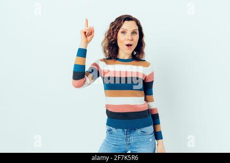 Fröhliche junge Frau mit lässigem gestreiftem Pullover und blauer Jeans Zeigt den Zeigefinger auf dem weißen Studiohintergrund an Mit überrascht und ha Stockfoto