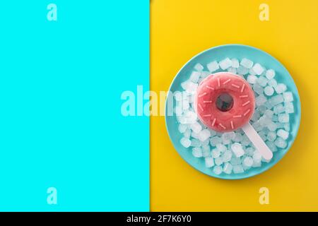 Erdbeerdonut-Popsicle und zerkleinertes Eis auf gelbem und blauem Hintergrund. Stockfoto