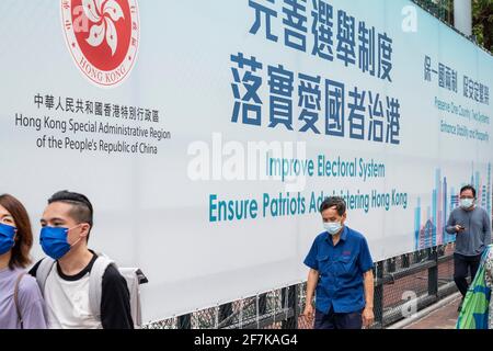 Hongkong, China. April 2021. Menschen mit Gesichtsmasken kommen an einem Transparent der Regierung vorbei, das an einem Zaun auf dem Spielplatz hängt und auf dem steht: „Verbessere das Wahlsystem, Sorge dafür, dass Patrioten Hongkong verwalten“. (Foto von Miguel Candela/SOPA Images/Sipa USA) Quelle: SIPA USA/Alamy Live News Stockfoto