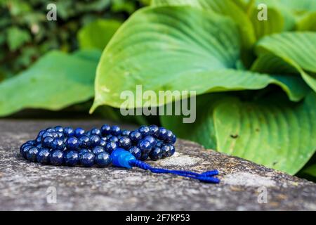 Blaue mala Perlen (Lapislazuli) auf Stein mit grünen Blättern im Hintergrund. Yoga- und Meditationszubehör. Stockfoto