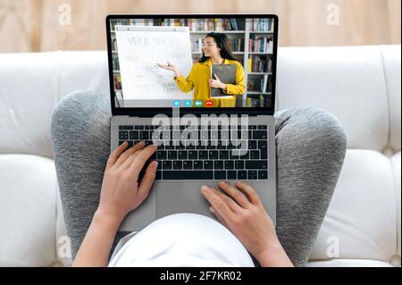Online-Training während der Schwangerschaft. Eine Schwangere hält einen Laptop, während sie auf der Couch sitzt, hört einen Online-Vortrag, auf dem Laptop-Bildschirm zeigt eine clevere Lehrerin Informationen während eines Videoanrufs Stockfoto
