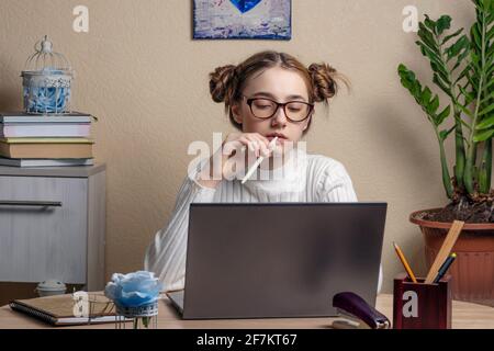 Schöne ernsthafte Teenager-Mädchen in Brille ist online auf einem Laptop verlobt. Ein Teenager-Mädchen schaut nachdenklich auf einen Laptop-Bildschirm und kaut auf einem Stift. Onli Stockfoto