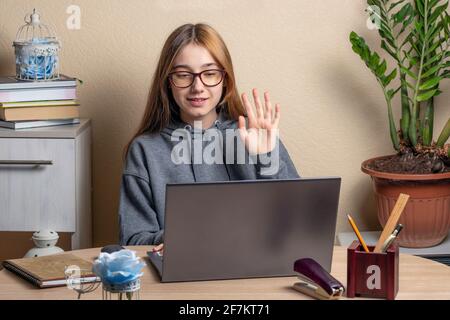 Schönes Teenager-Mädchen mit Brille sitzt vor Laptops. Das Teenager-Mädchen winkt mit der Hand auf den Laptop-Bildschirm. Fernkommunikationskonzept mit Frie Stockfoto