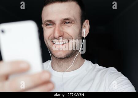 Junger bärtiger Kerl, der mit Kopfhörern in das Telefon lächelt Ein weißes T-Shirt auf dunklem Hintergrund Stockfoto