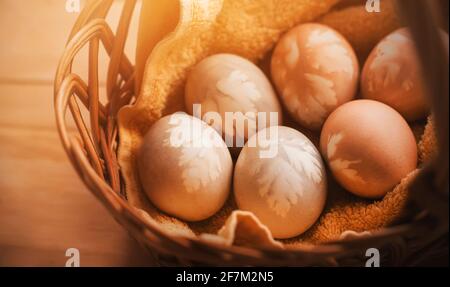 Hühnereier, die mit natürlichen Farbstoffen mit Blattmustern bemalt sind, liegen in einem Weidenkorb mit einem gelben Handtuch, das vom Sonnenlicht beleuchtet wird. Osterdeko Stockfoto