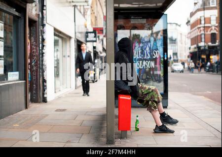 Junger Mann im Kapuzenpullover sitzt in der Bushaltestelle und wartet auf den Bus. Alltag in der Stadt. Bethnal Green Road, East London, Großbritannien. Juli 2015 Stockfoto