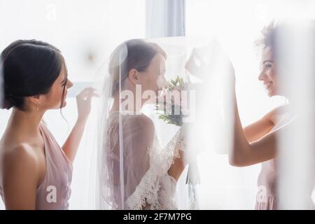 Seitenansicht der glücklichen interracial Brautjungfern in der Nähe lächelnde Braut in Schleier auf verschwommenem Vordergrund Stockfoto