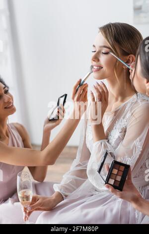 Multikulturelle Brautjungfern, die Lidschatten und Gesichtspuder auf Pretty auftragen Braut im Schlafzimmer Stockfoto